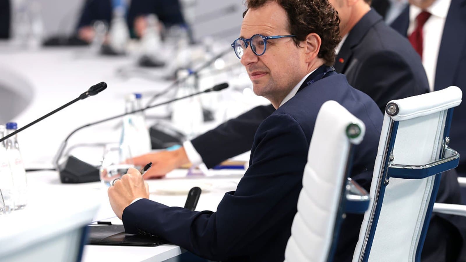 A man in a navy suit and round glasses seated at a long conference table with microphones, turning towards the camera. Foto d'archivio.