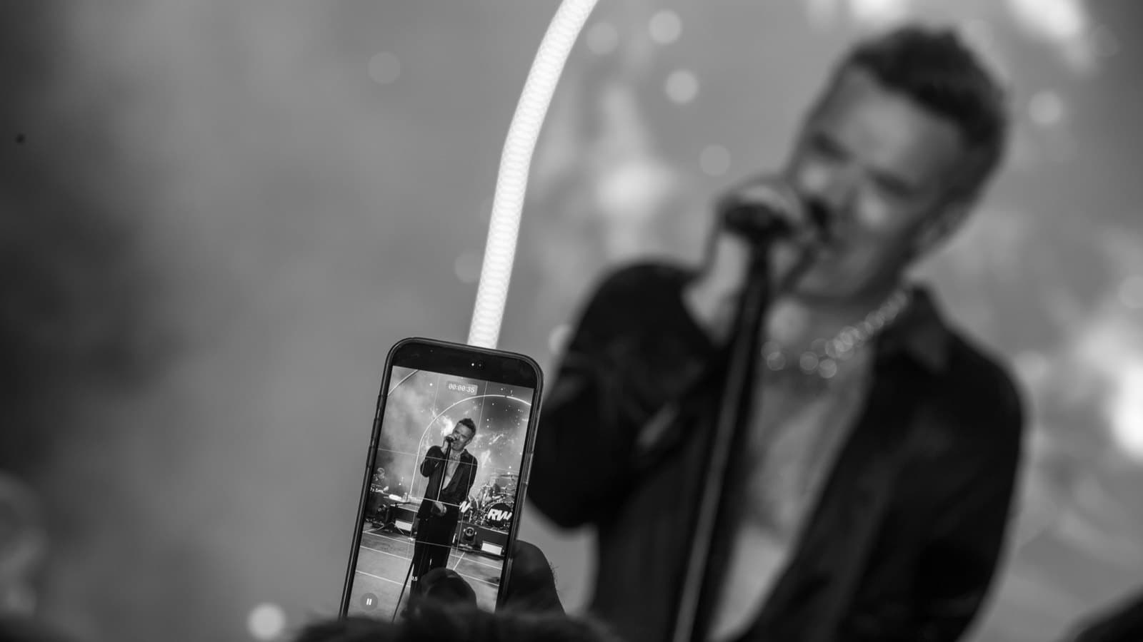 A black and white photograph of Robbie Williams singing at a microphone stand, seen past a phone screen in the crowd filming him. Foto d'archivio.