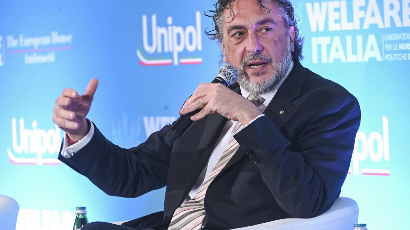 Carlo Cimbri, chairman of Unipol, speaking into a handheld microphone on a conference stage in front of a blue Unipol backdrop.