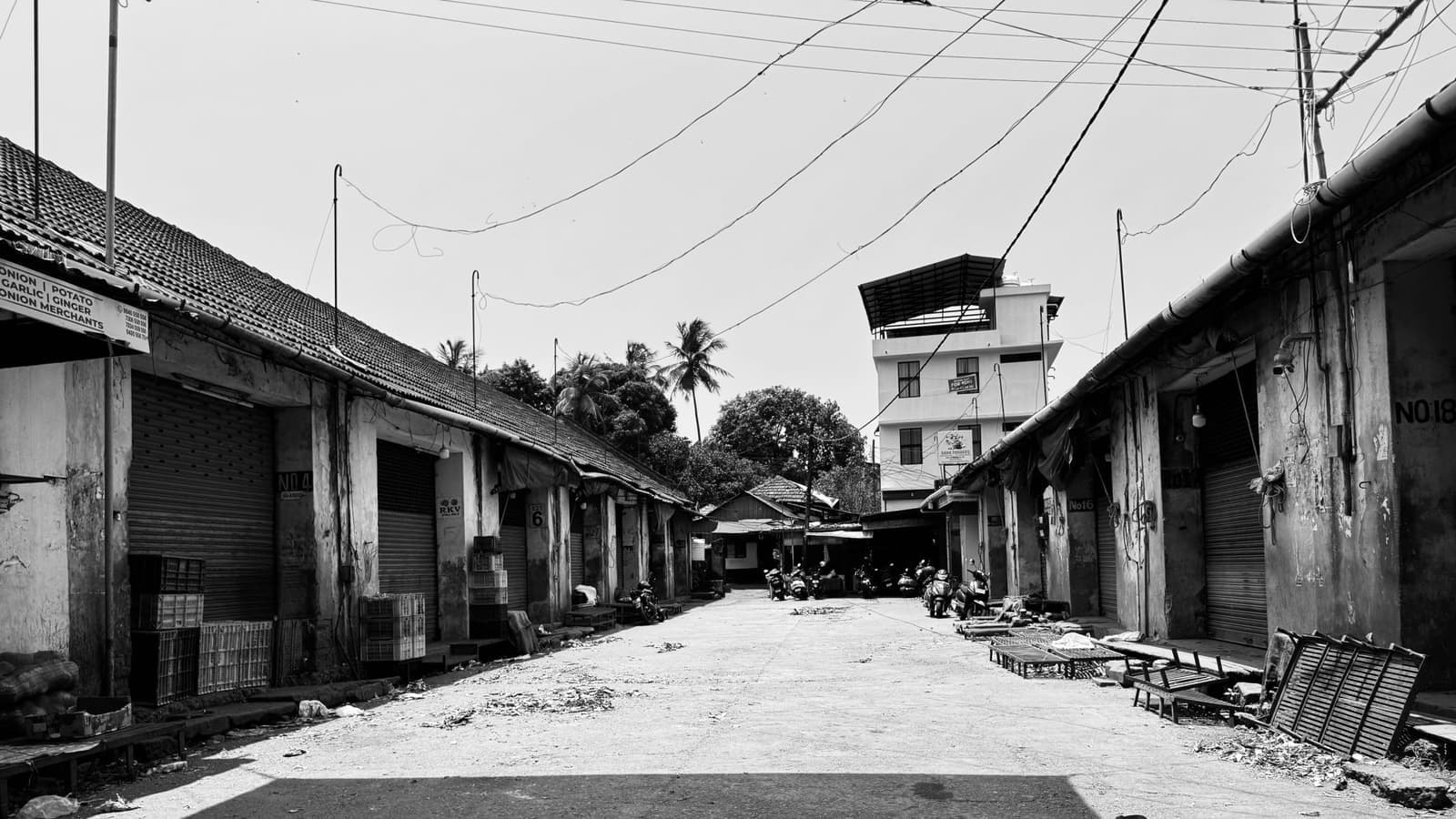 A black and white photo of a deserted market street featuring several closed shop fronts with rolling metal shutters, crates stacked outside, and scattered debris on the ground. Calicut old vegetable market. Foto d'archivio.