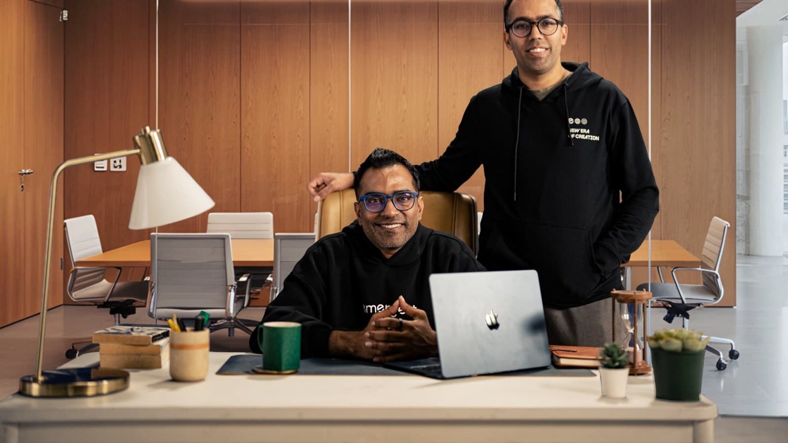 Two company founders in a meeting room, one seated at a desk with a laptop and one standing beside him. Company photograph. Foto d'archivio.