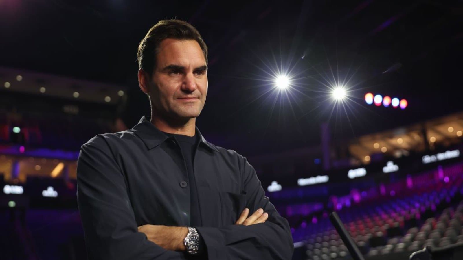 Roger Federer standing with his arms folded courtside in an empty arena lit in purple and blue. Foto d'archivio.