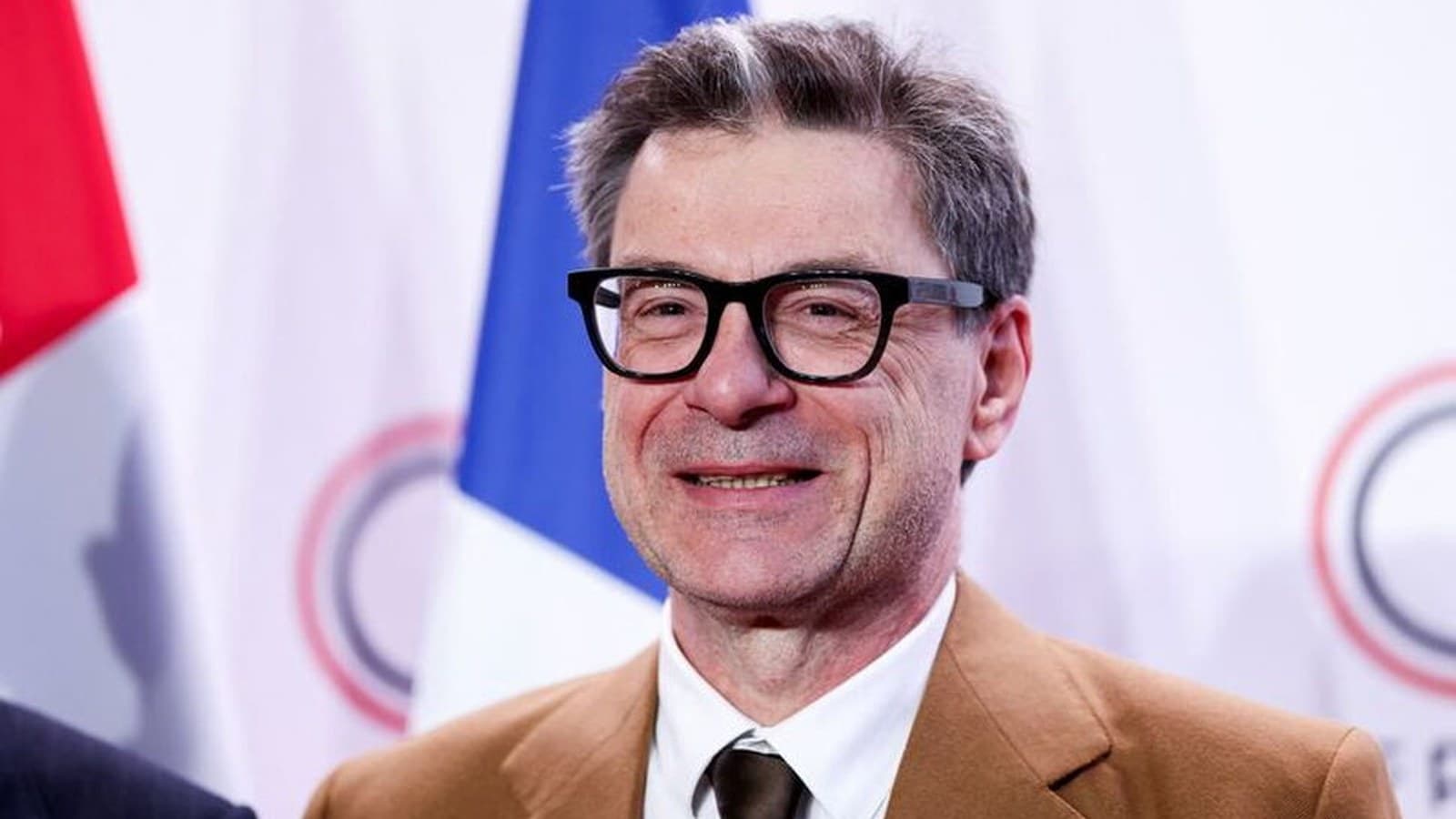 Giancarlo Giorgetti sorride in giacca cammello davanti a bandiere europee.