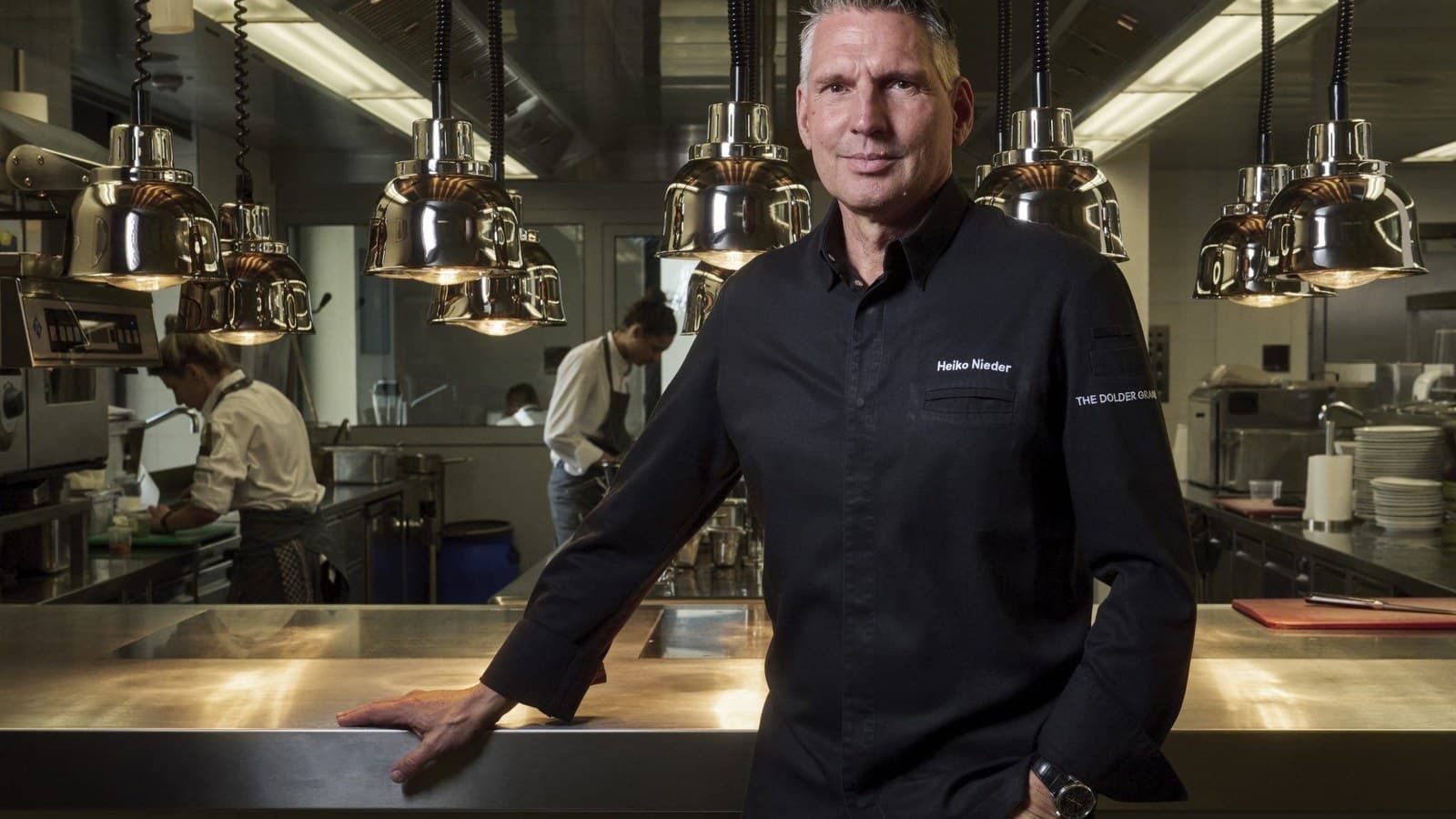 Lo chef Heiko Nieder in piedi nella cucina del suo ristorante sotto file di lampade in rame appese, con una mano appoggiata sul bancone in acciaio.