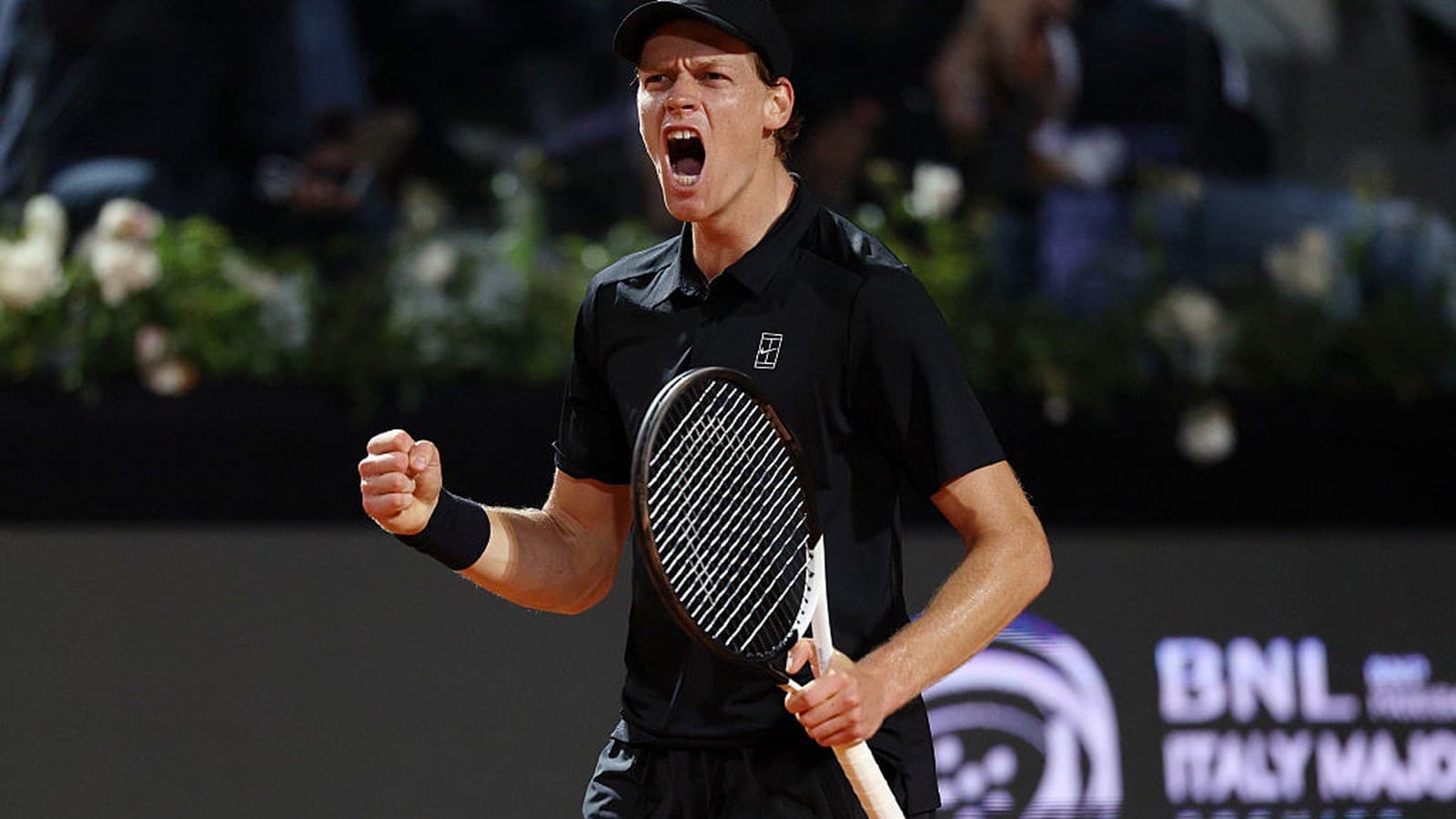 Jannik Sinner stringe il pugno ed esulta urlando su un campo da tennis, con cappellino e maglia nera.