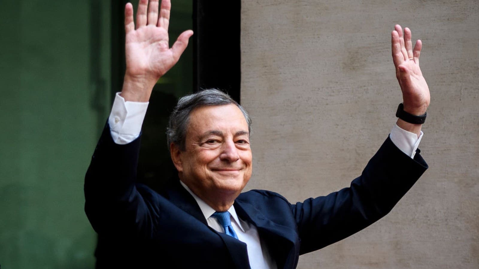 Mario Draghi, in abito scuro e cravatta blu, saluta con entrambe le mani alzate, sorridendo.