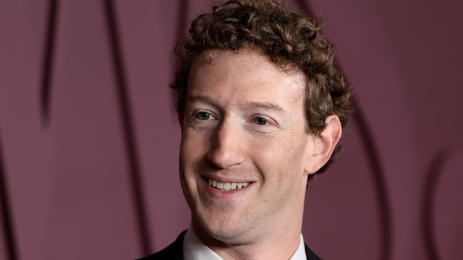 Mark Zuckerberg smiles in a dark suit against a maroon patterned backdrop.