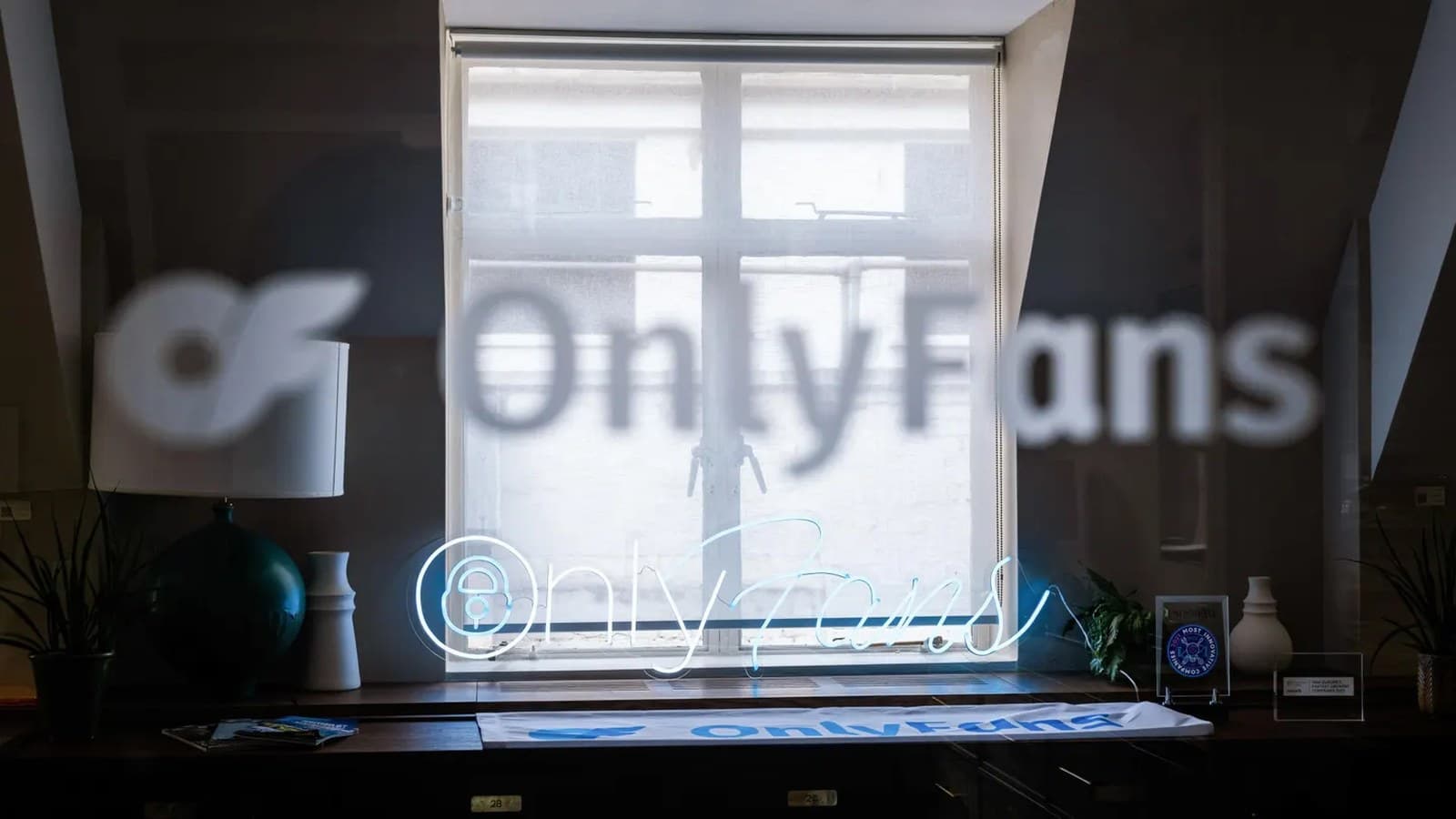 Il logo di OnlyFans si illumina in blu al neon su una scrivania davanti a una finestra, con il nome proiettato sulla parete dietro.