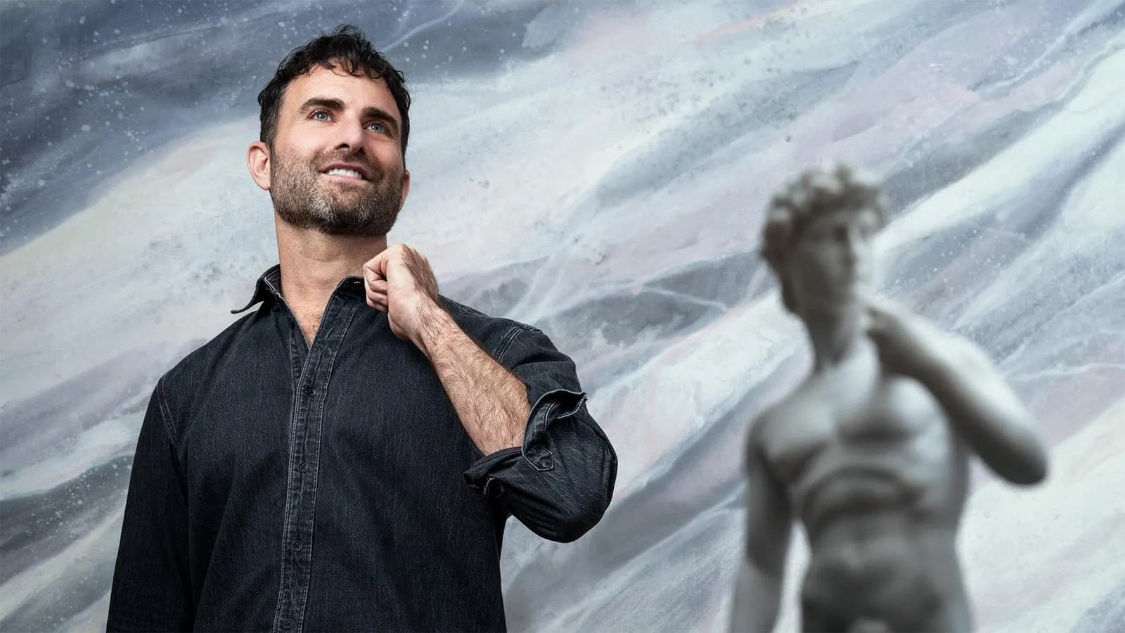 Peter Rahal sorride sistemandosi il colletto davanti a uno sfondo artistico con una statua sfocata del David.