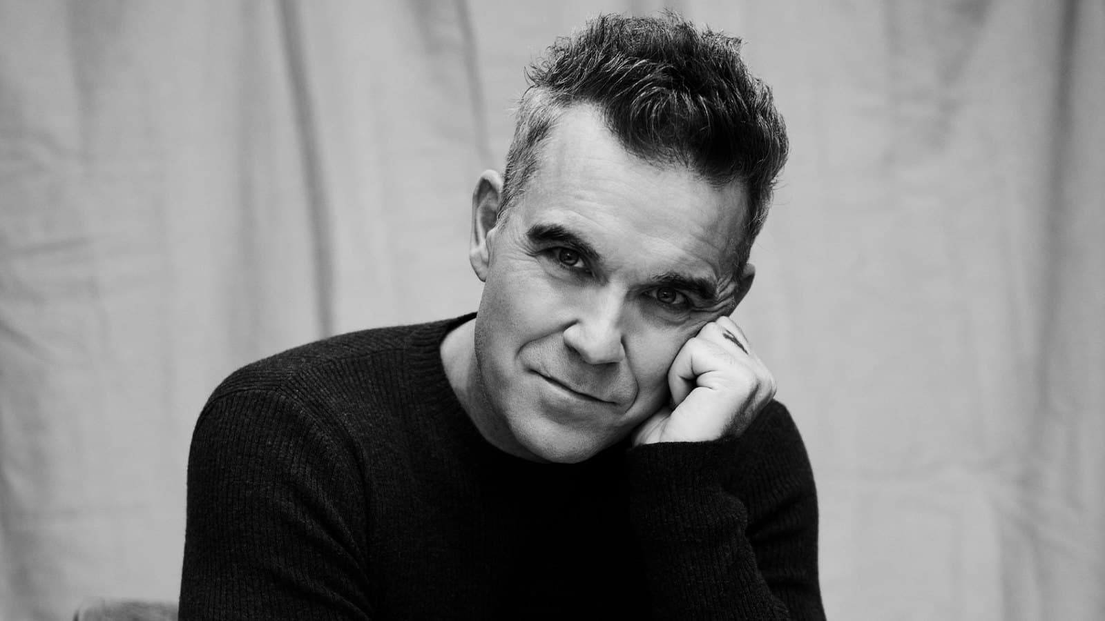 Robbie Williams in a black suit against a black background, looking towards the camera. Foto d'archivio.