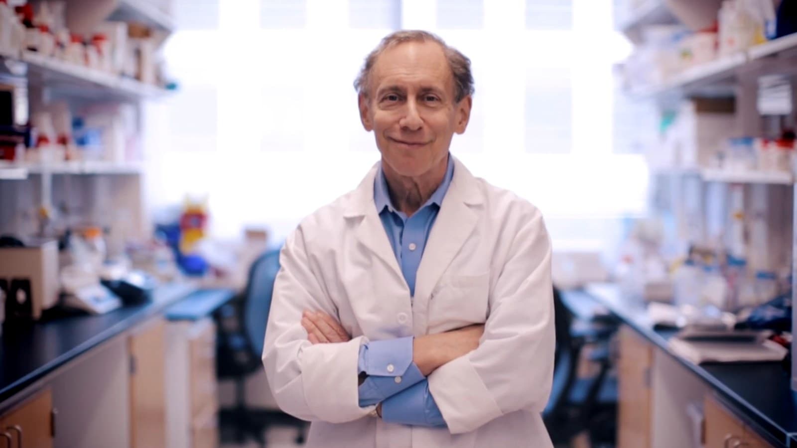 Robert Langer, in camice bianco e braccia conserte, sorride in un laboratorio.