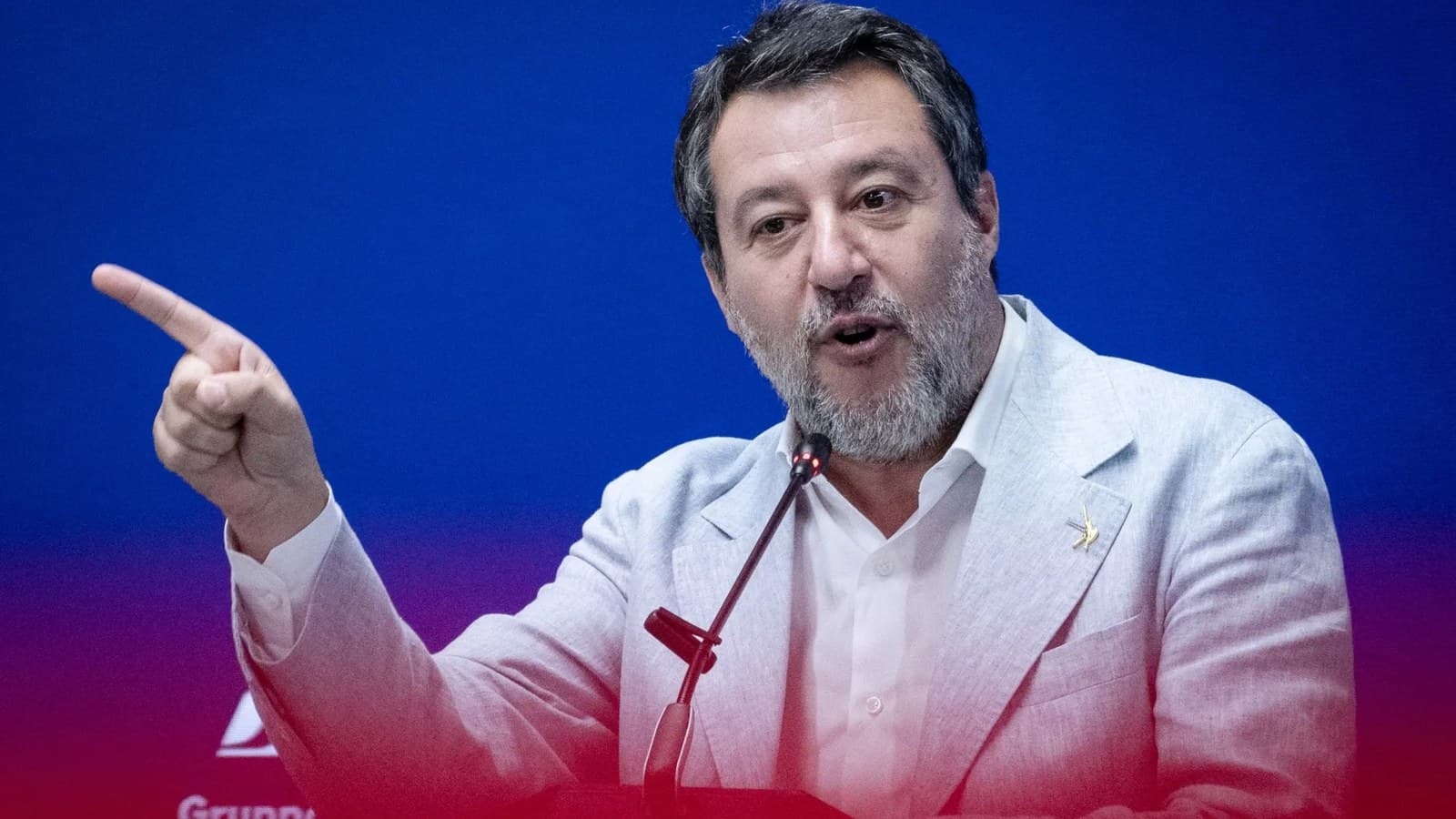 Matteo Salvini gestures while speaking at a podium against a blue and red backdrop.