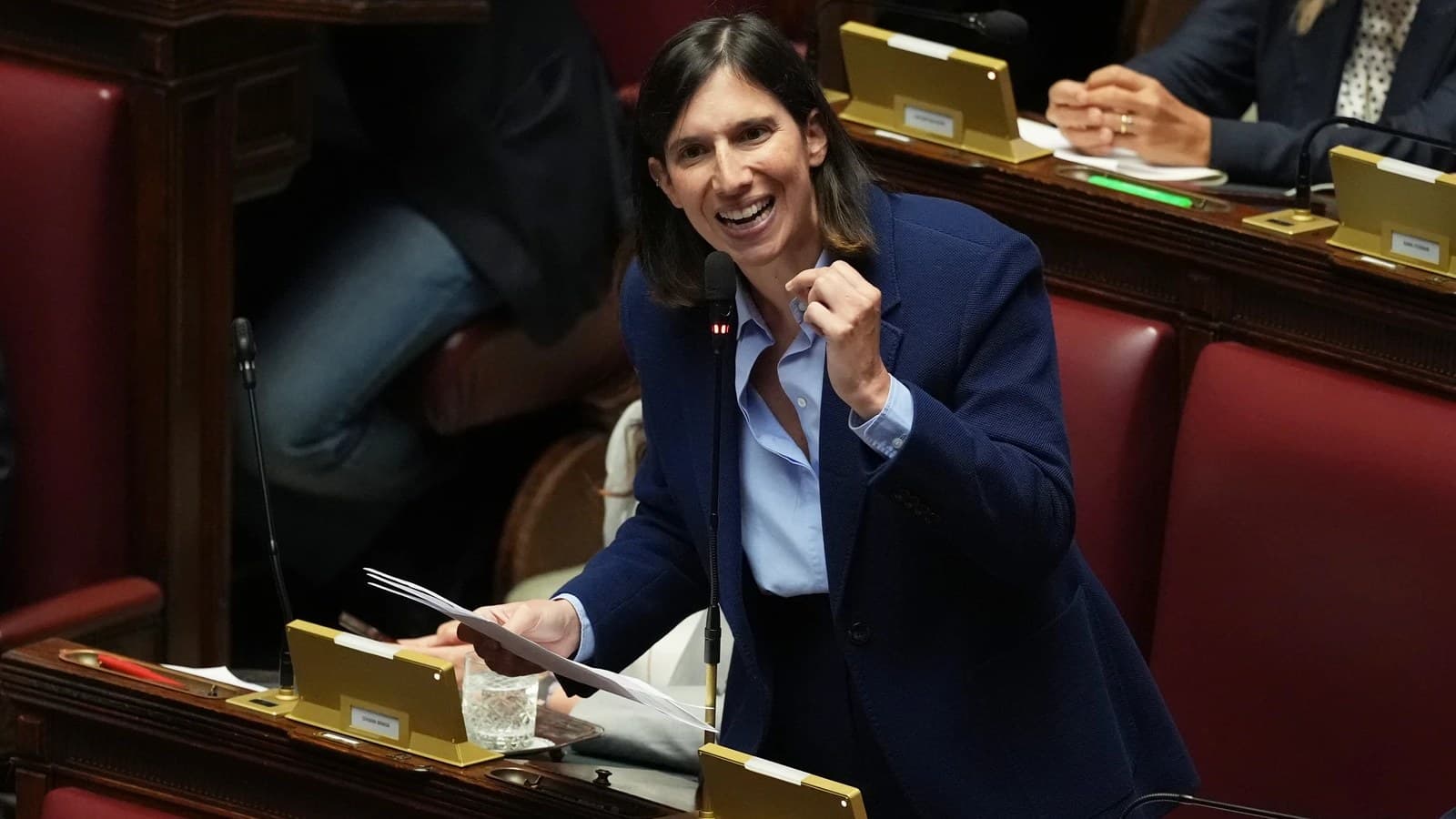 Elly Schlein sorride e gesticola mentre parla da un leggio alla Camera dei deputati.