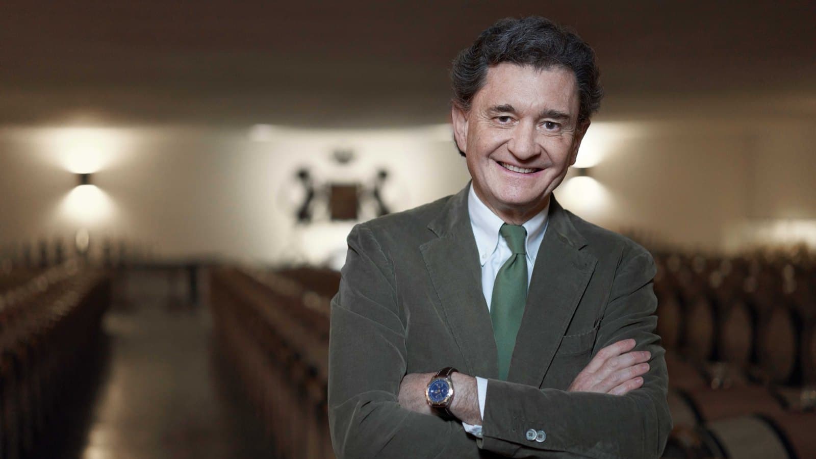 A man in a green corduroy jacket and green tie standing with his arms folded in a barrel hall, rows of oak barrels receding behind him.