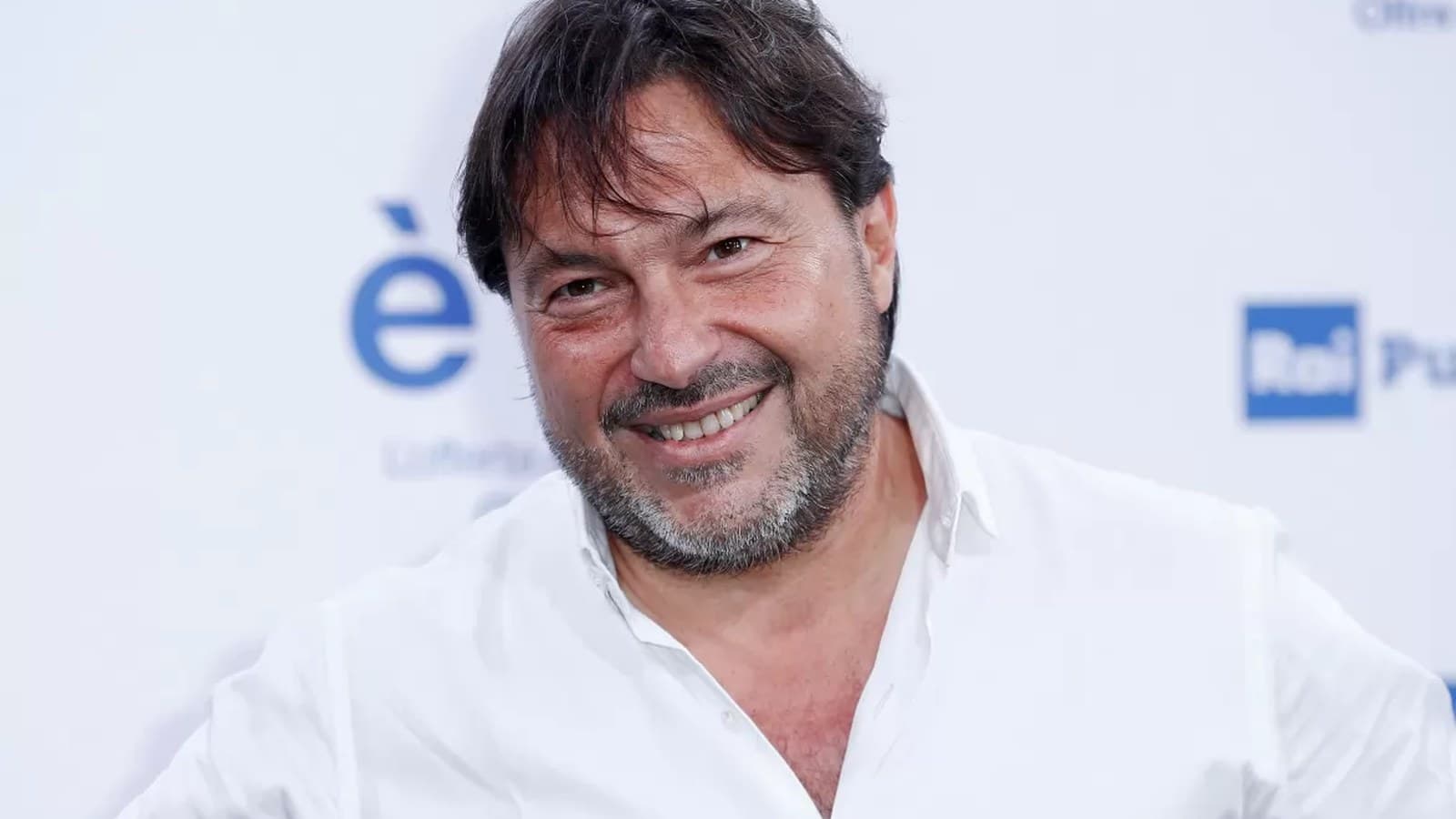 Sigfrido Ranucci sorride in camicia bianca davanti a uno sfondo con il logo Rai.