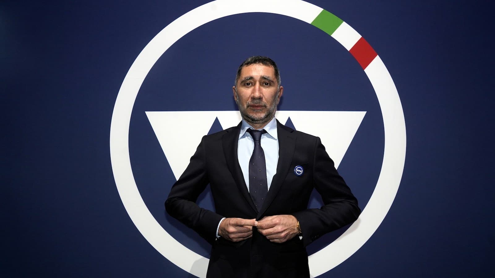 Cristiano Crippa, president of Technoprobe, stands in a dark suit in front of a large circular company logo in navy and white.