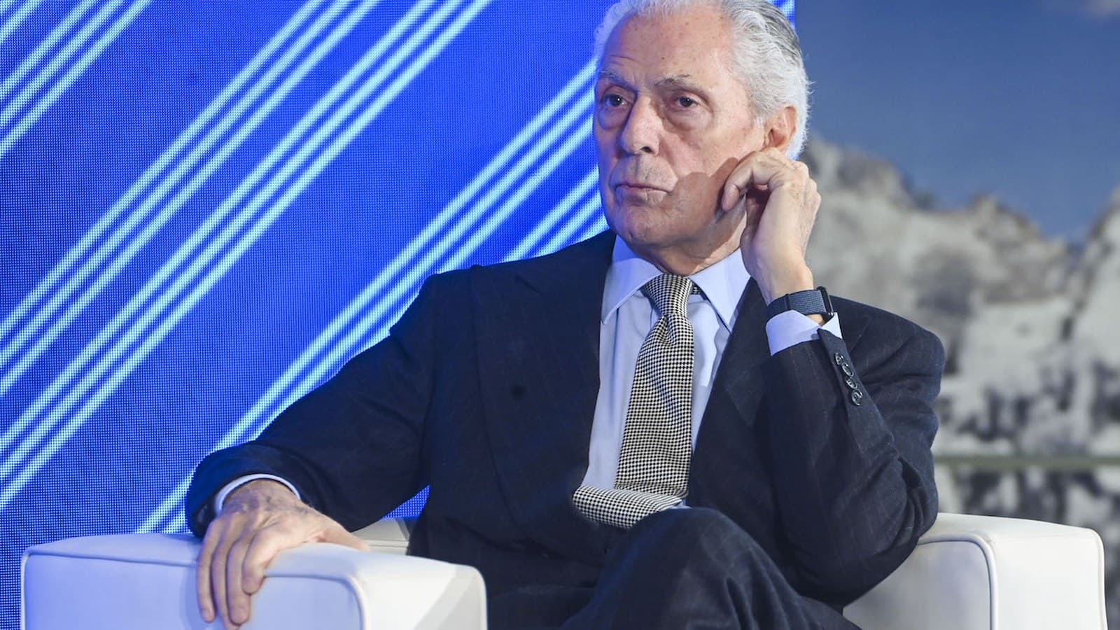 Marco Tronchetti Provera sits in a white armchair on stage, resting his hand near his face.