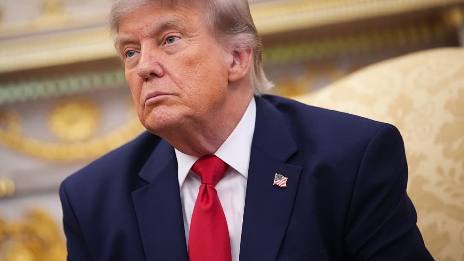 Donald Trump in a navy suit and red tie, seated in the Oval Office and looking off camera.