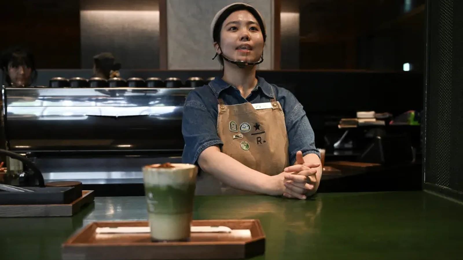 Una barista in grembiule si trova dietro al bancone con una bevanda al matcha, parlando con qualcuno fuori campo.