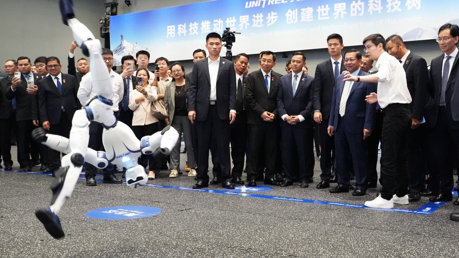 A white humanoid robot performs an acrobatic flip on stage as a crowd of officials and photographers watches at a Unitree event.