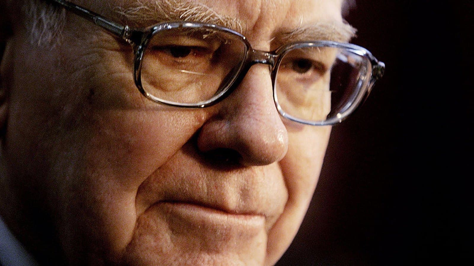A close portrait of Warren Buffett in glasses, lit from one side against a dark background.