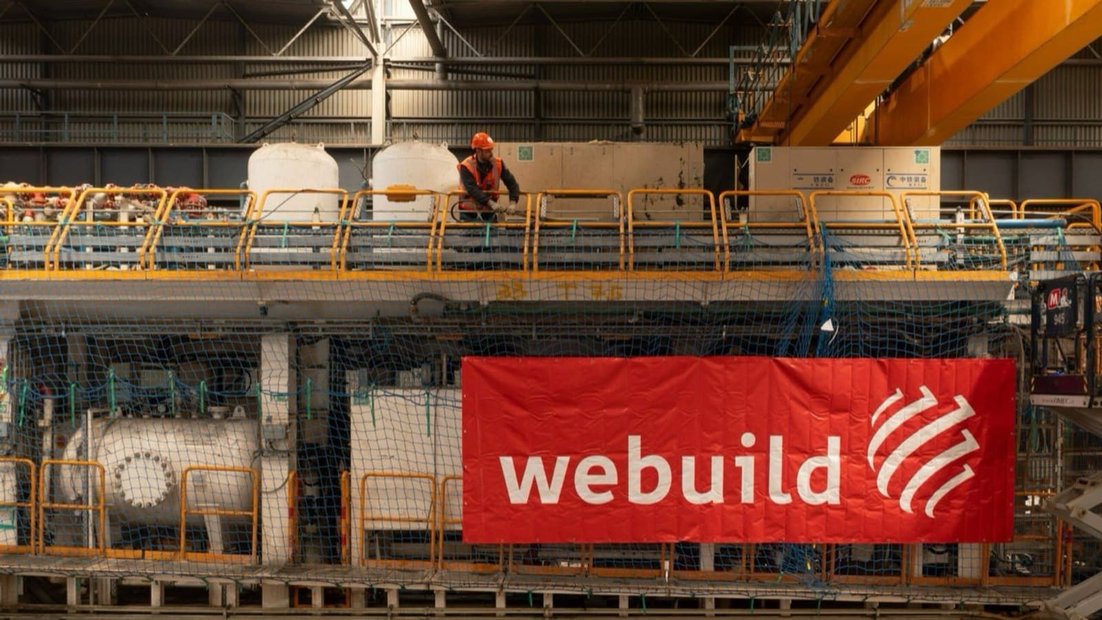 A worker on a tunnel boring machine in an assembly hall, beside a red Webuild banner. File photograph. Foto d'archivio.