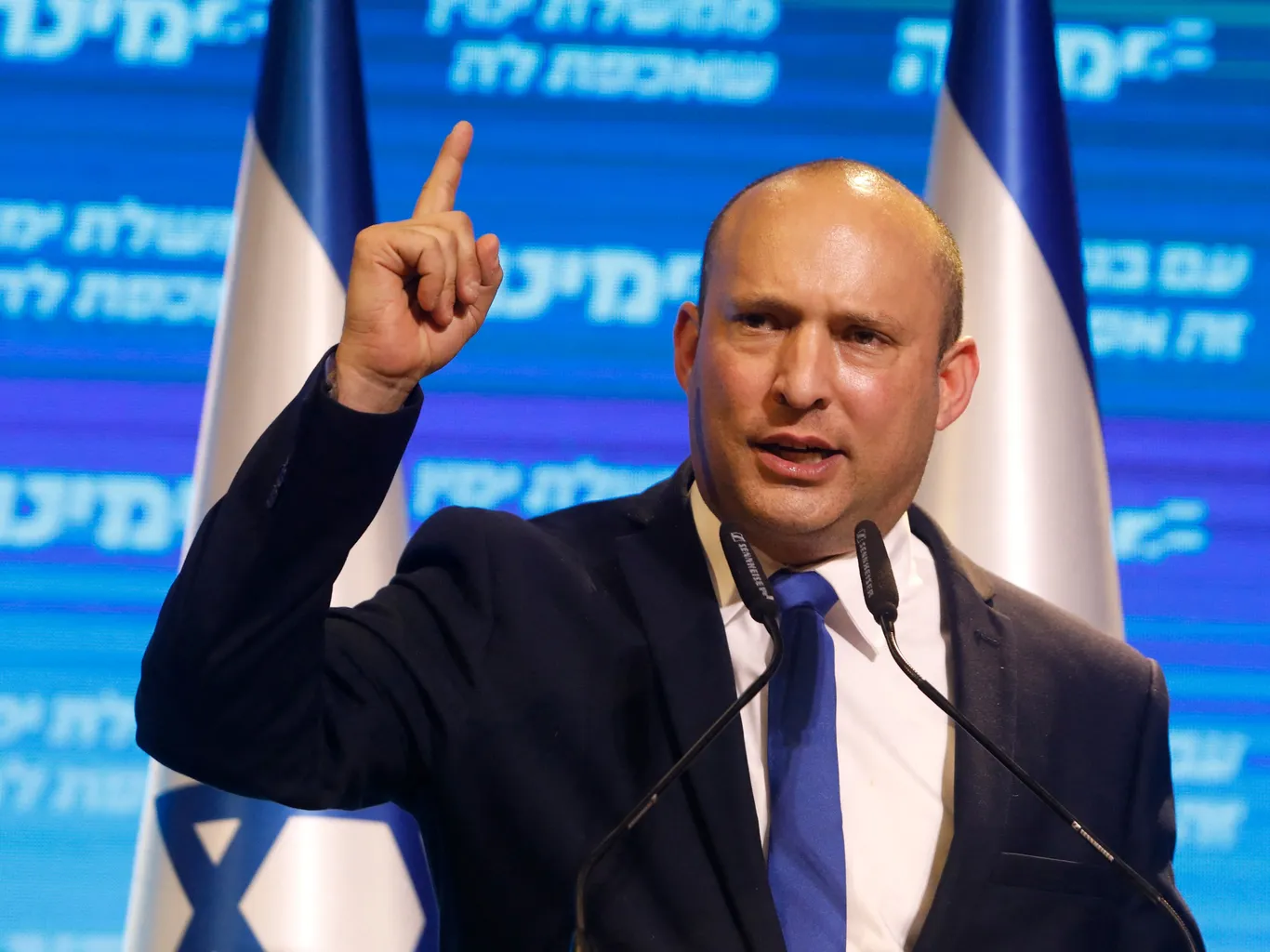 Naftali Bennett gesticola mentre parla a un podio davanti alle bandiere israeliane.