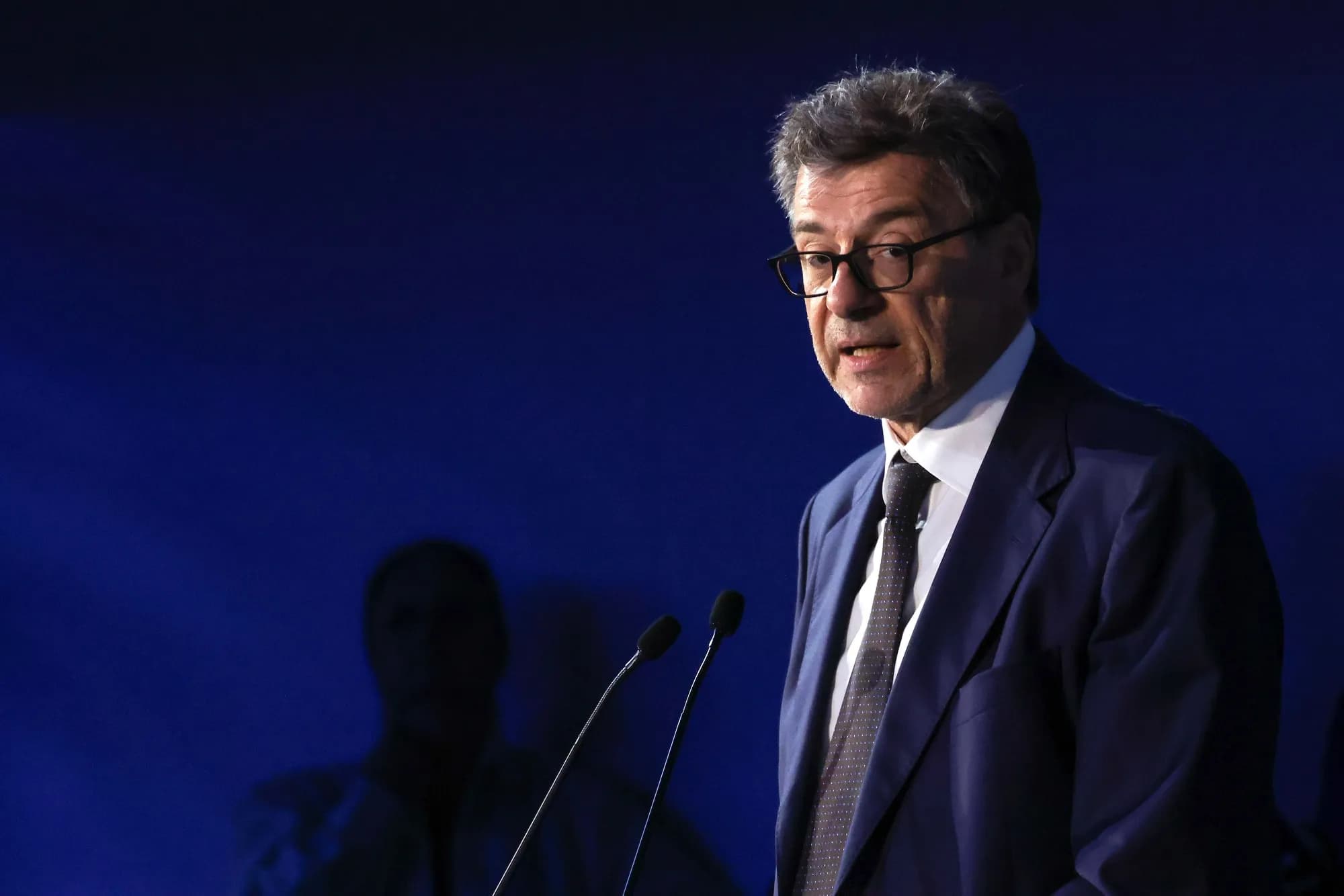 Giancarlo Giorgetti parla a un podio con due microfoni davanti a uno sfondo blu scuro.
