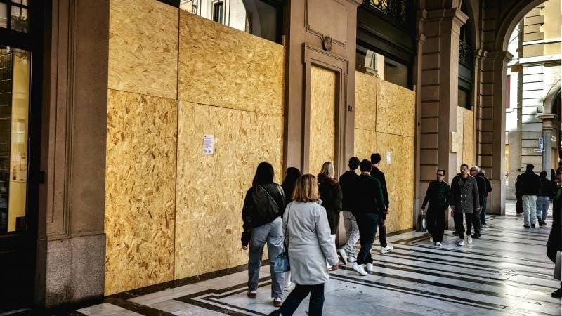 Pedoni camminano davanti a vetrine di negozi tappate con assi di legno sotto un portico in una città italiana.