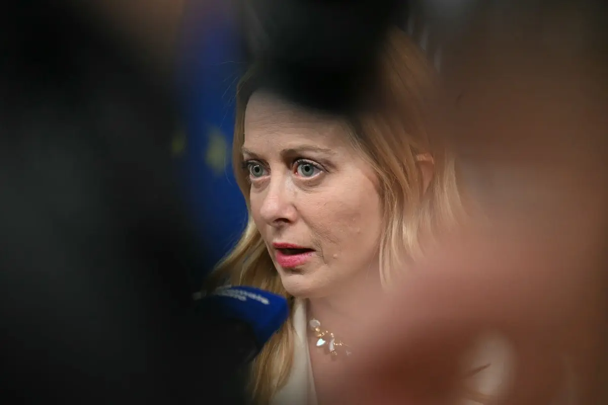 Giorgia Meloni parla ai giornalisti che tengono microfoni, con una bandiera dell'Unione europea visibile sullo sfondo.