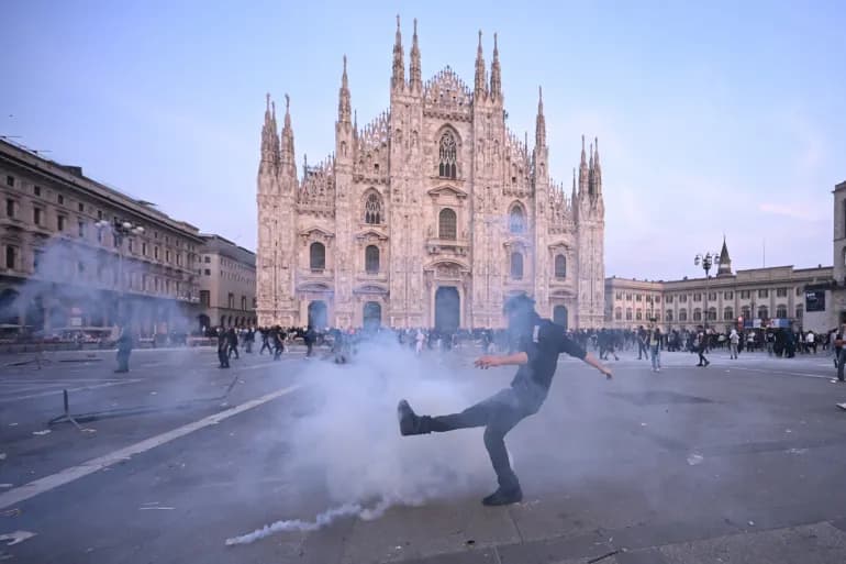 Un manifestante calcia un candelotto lacrimogeno davanti al Duomo di Milano mentre il fumo invade la piazza.
