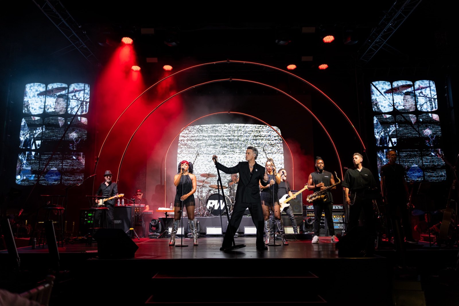 Veduta ampia del palco con Robbie Williams al centro, la band e le coriste attorno a lui sotto archi rossi illuminati.