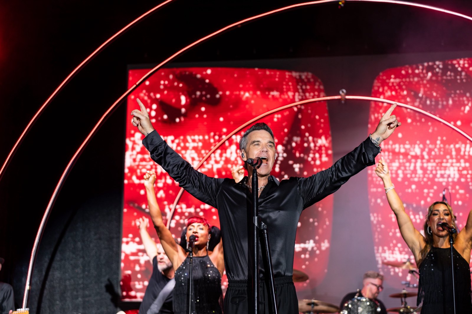 Robbie Williams sul palco con entrambe le braccia alzate e gli indici puntati verso l'alto, le coriste alle spalle su uno schermo rosso.