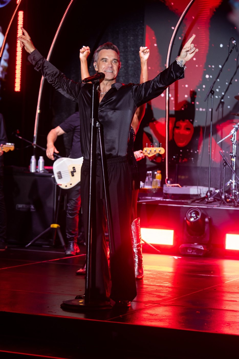 Robbie Williams all'asta del microfono, vestito di nero e con le braccia alzate, il palco illuminato di rosso alle spalle.