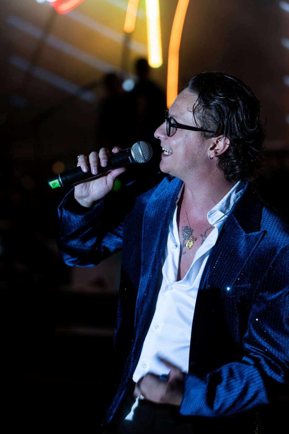 Un artista con giacca blu di paillettes canta al microfono sotto luci calde da palco.
