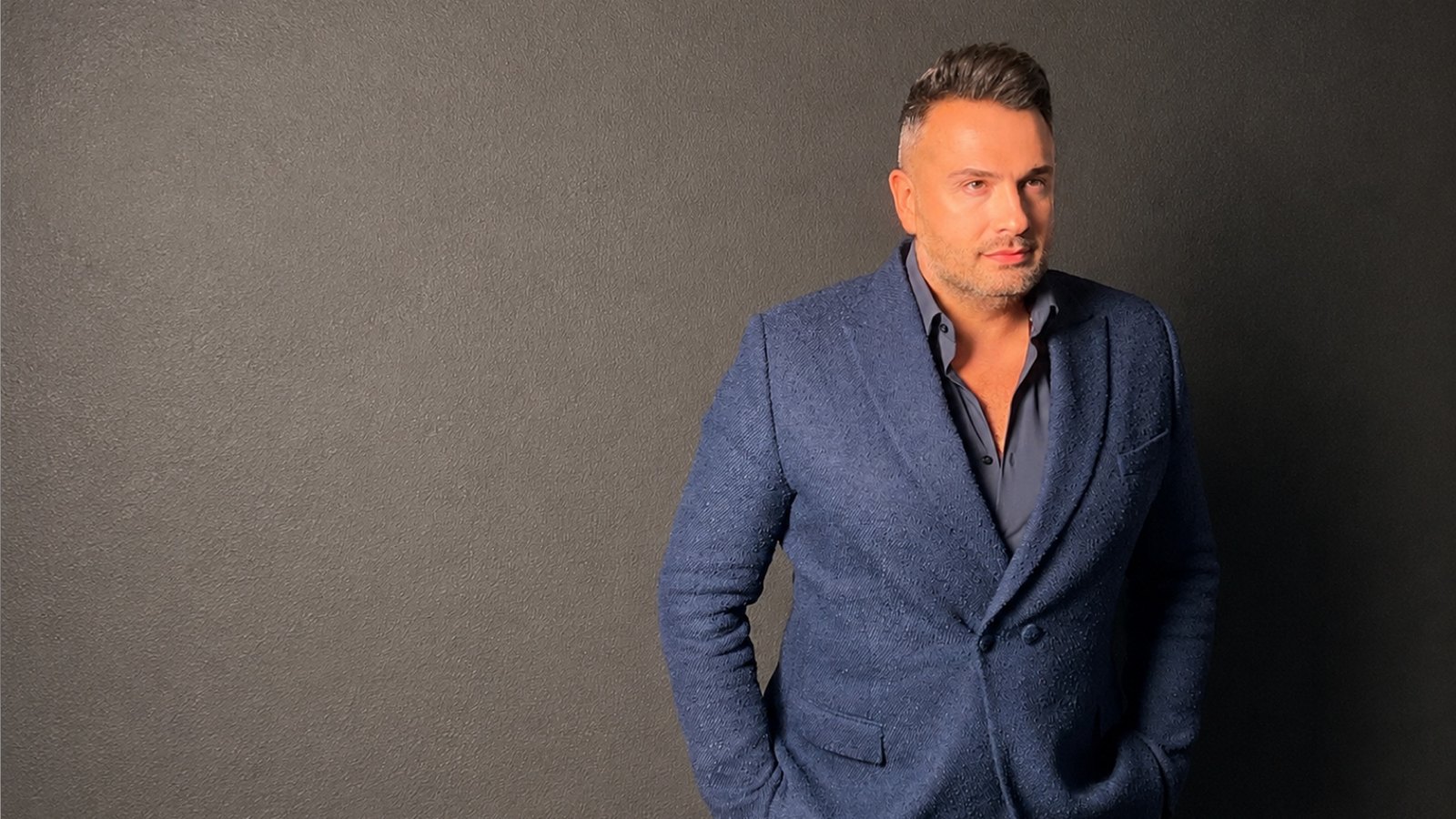 Maxim Berin in piedi davanti a una parete grigia, con una giacca blu in tessuto lavorato.
