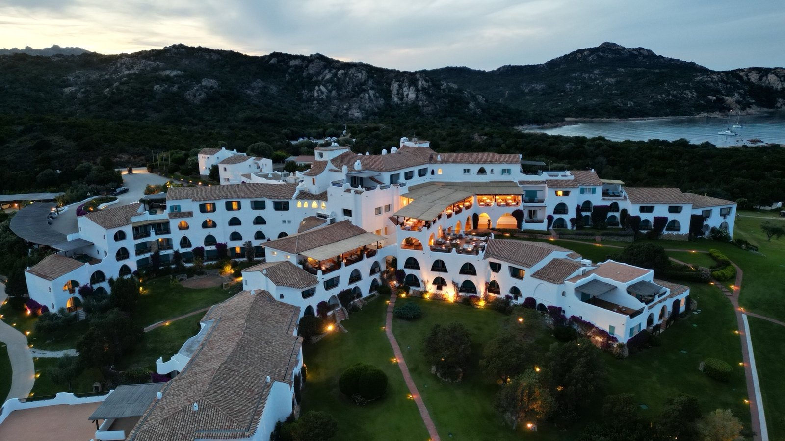 Veduta aerea al crepuscolo del Romazzino, A Belmond Hotel, con gli edifici bianchi illuminati tra le colline e il mare della Costa Smeralda.